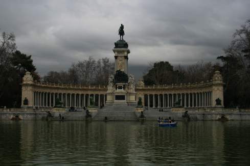 a statue in front of a pond with Buen Retiro Park in the background